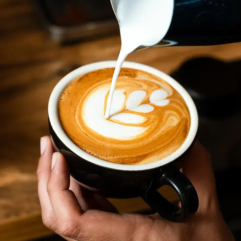 Leche vertida desde una jarra sobre un café con dibujo de hojas en la espuma, en taza negra sostenida con la mano