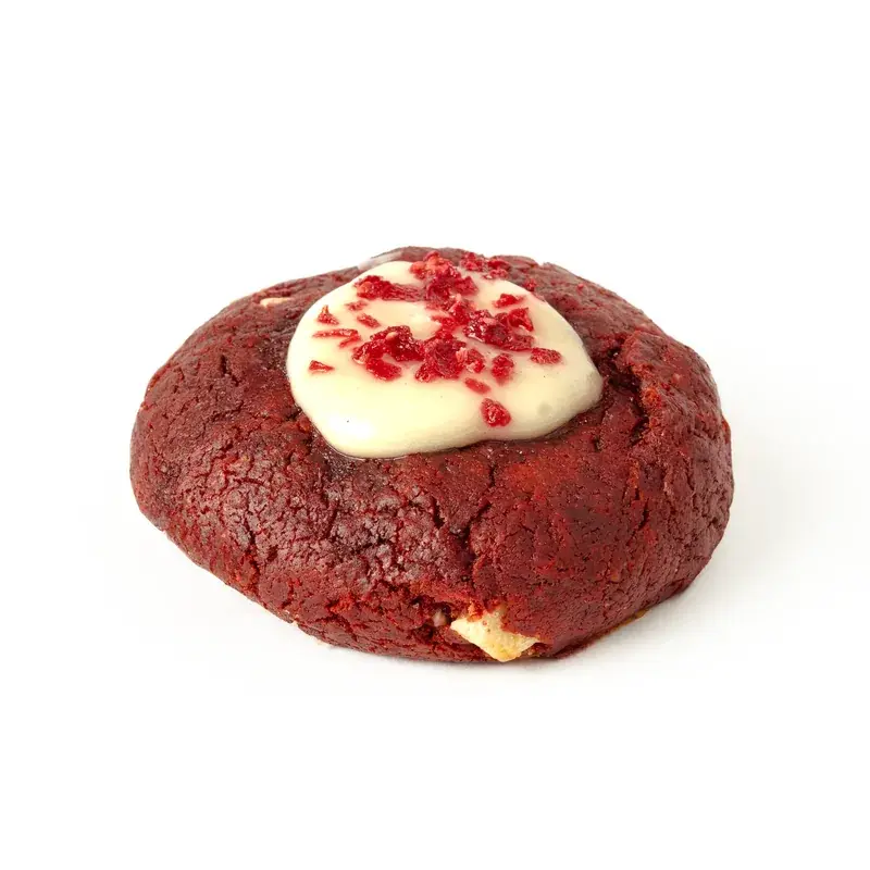 Cookie red velvet de masa roja con crema blanca y trocitos rojos por encima, sobre fondo blanco