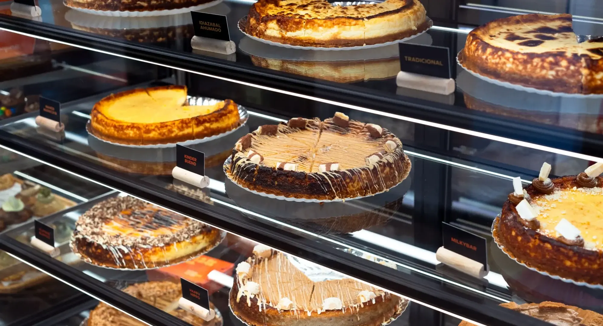 Vitrina de una tienda Cheesequería con tartas de queso enteras en sus estantes y el cartel de cada sabor