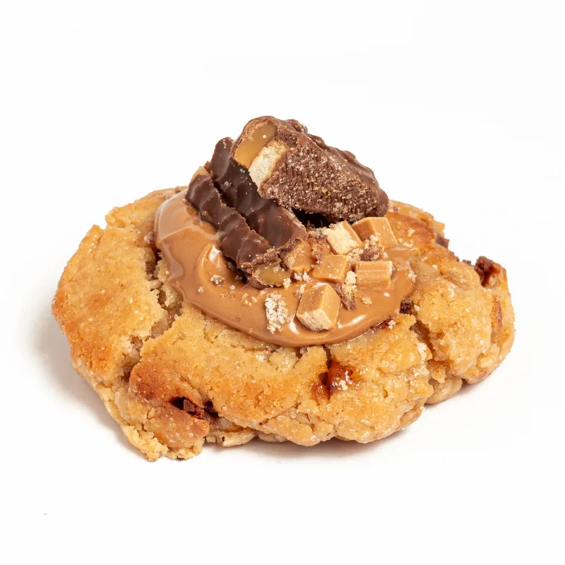 Cookie de masa dorada con crema de caramelo, trozos de Twix y dados de caramelo por encima, sobre fondo blanco